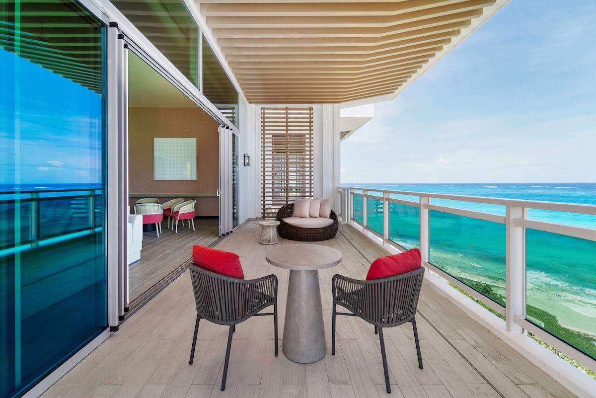 Resort suite balcony with two red-cushioned chairs facing turquoise ocean water, woven lounger, and shaded seating areas.