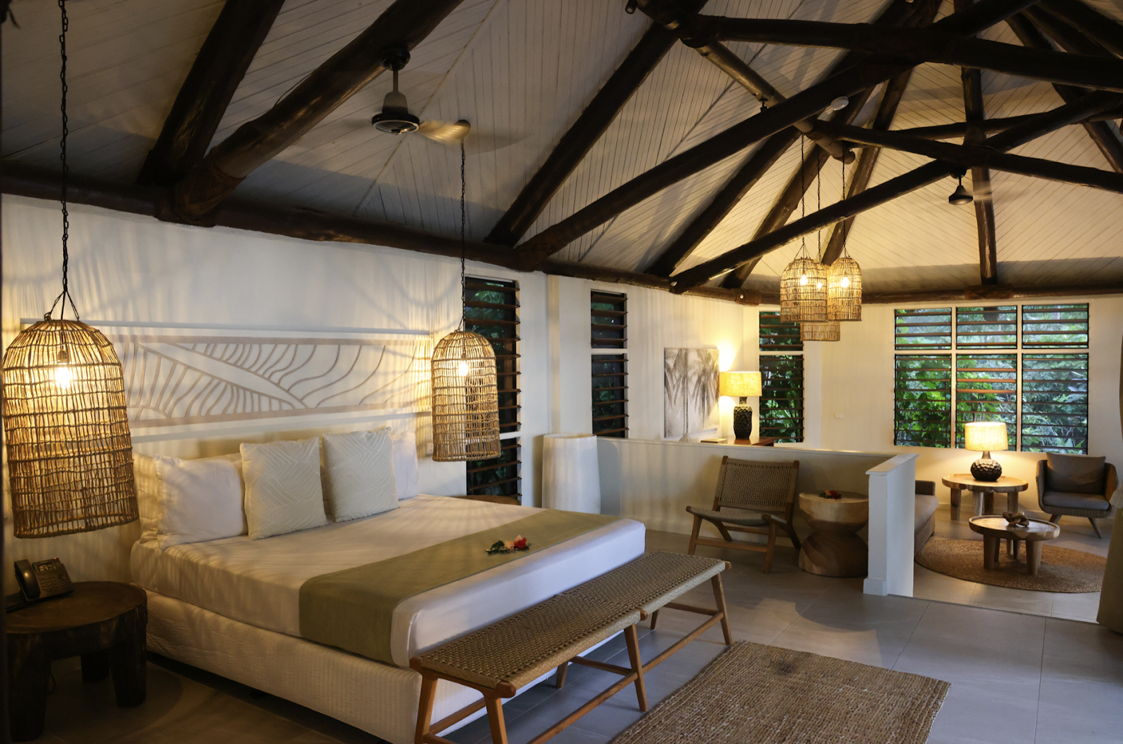 Spacious resort bedroom with woven pendant lights, wooden beam ceiling, king bed with cream linens, and lounge area with chairs overlooking tropical gardens through large windows.