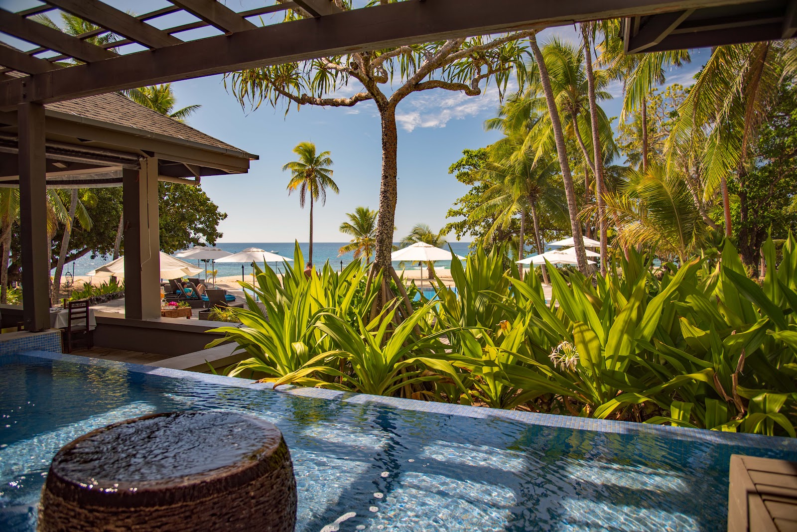 Pool with woven poufs in foreground overlooks tropical beach with palm trees, lounge chairs, and ocean beyond.