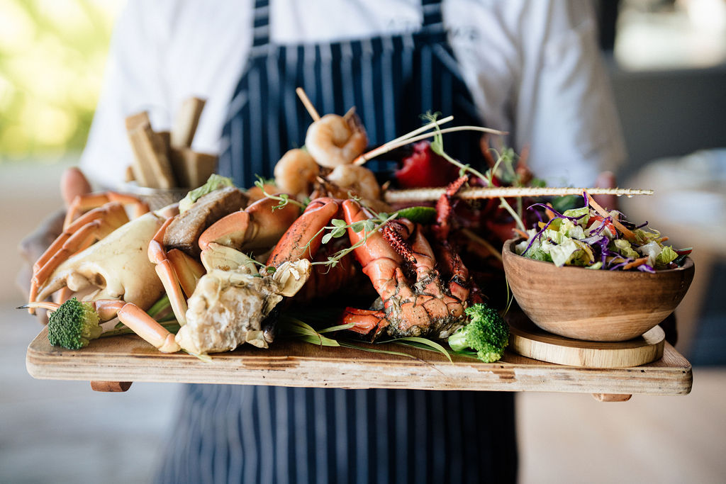 Chef in striped apron holding wooden board with grilled lobster, vegetables, and microgreens arranged for plating.