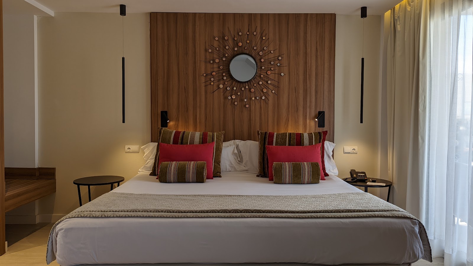 Modern resort suite with bed visible through wooden partition, sofa with yellow pillows, round ottoman, and large windows with cream curtains at Zafiro Palace Alcudia.