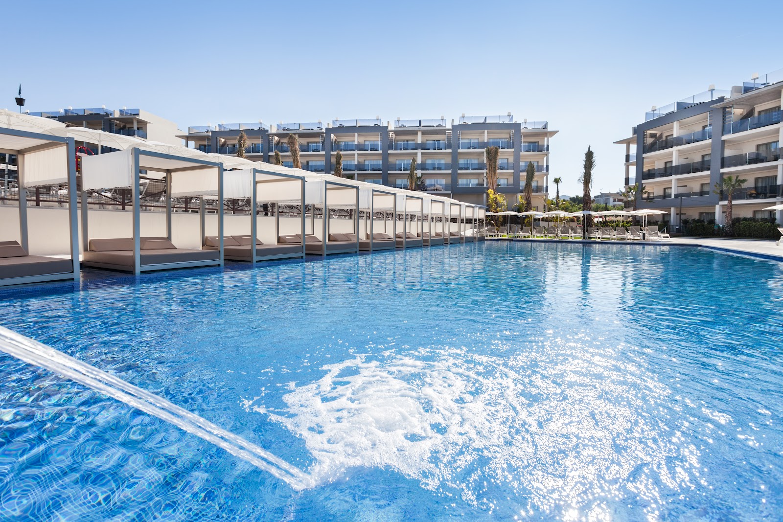 Large outdoor pool with white cabanas, loungers, and modern multi-story hotel buildings under clear blue sky at Zafiro Palace Alcudia.