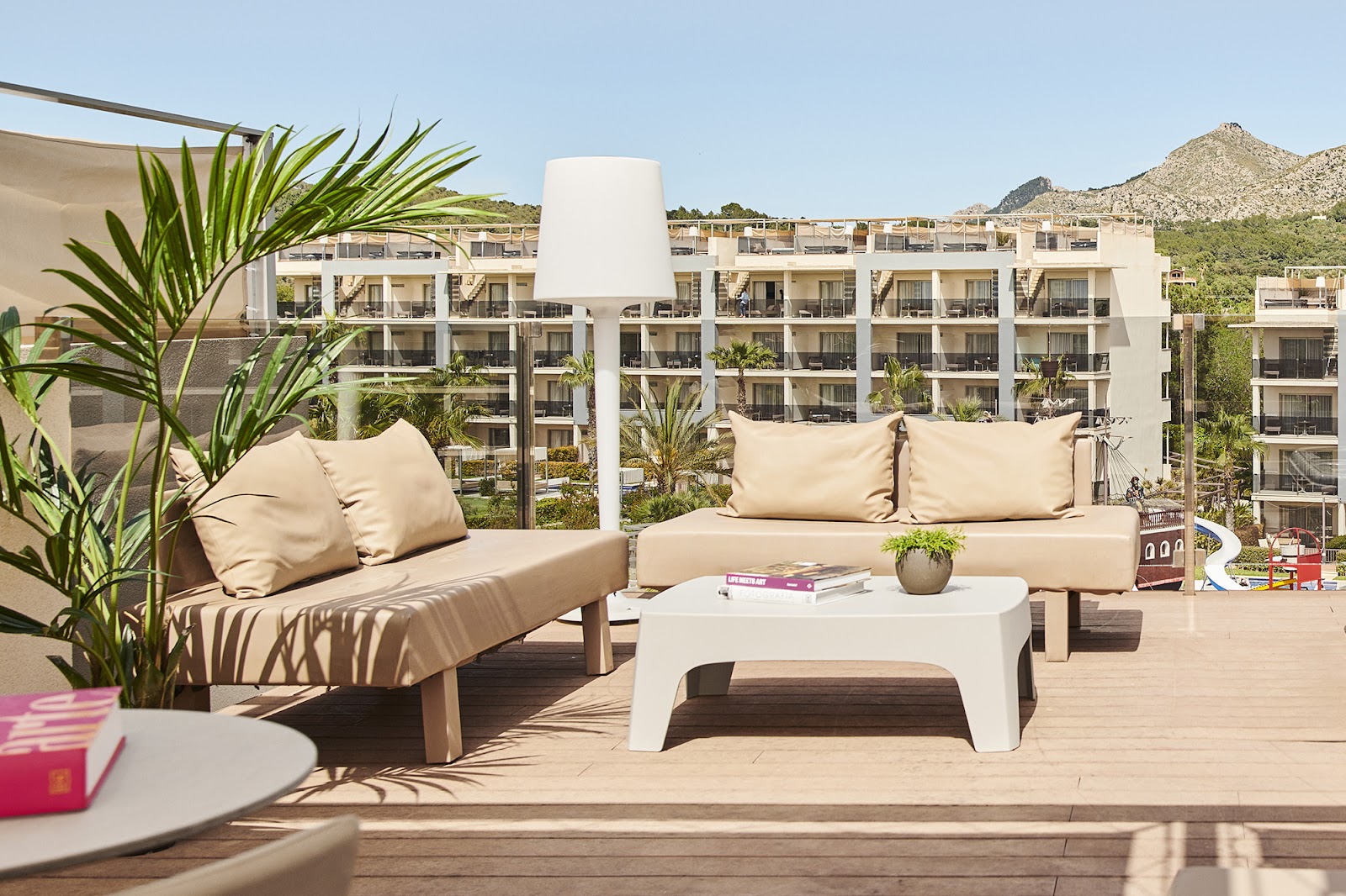 Lounge seating with cream cushions on a wooden terrace at Zafiro Palace Alcudia, with resort buildings and rocky hills beyond.