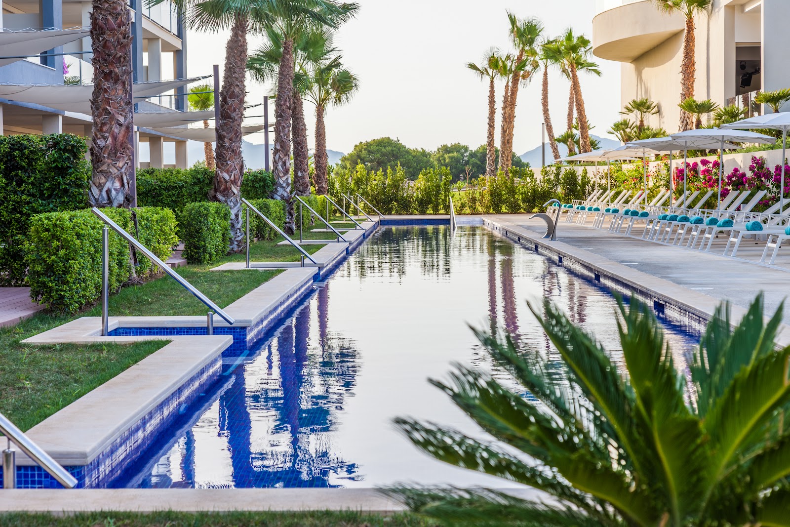 Zafiro Palace Alcudia's lagoon-style pool with blue-tiled edges reflects tall palms and modern white buildings, lined with lounge chairs and umbrellas.