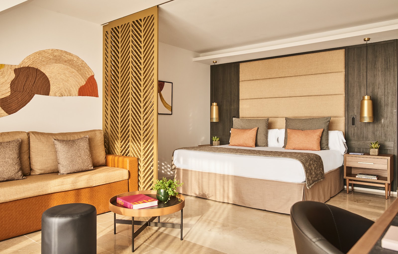 Resort suite with king bed, tan upholstered headboard, sitting area with gold sofa, geometric room divider, and abstract wall art in warm tones.
