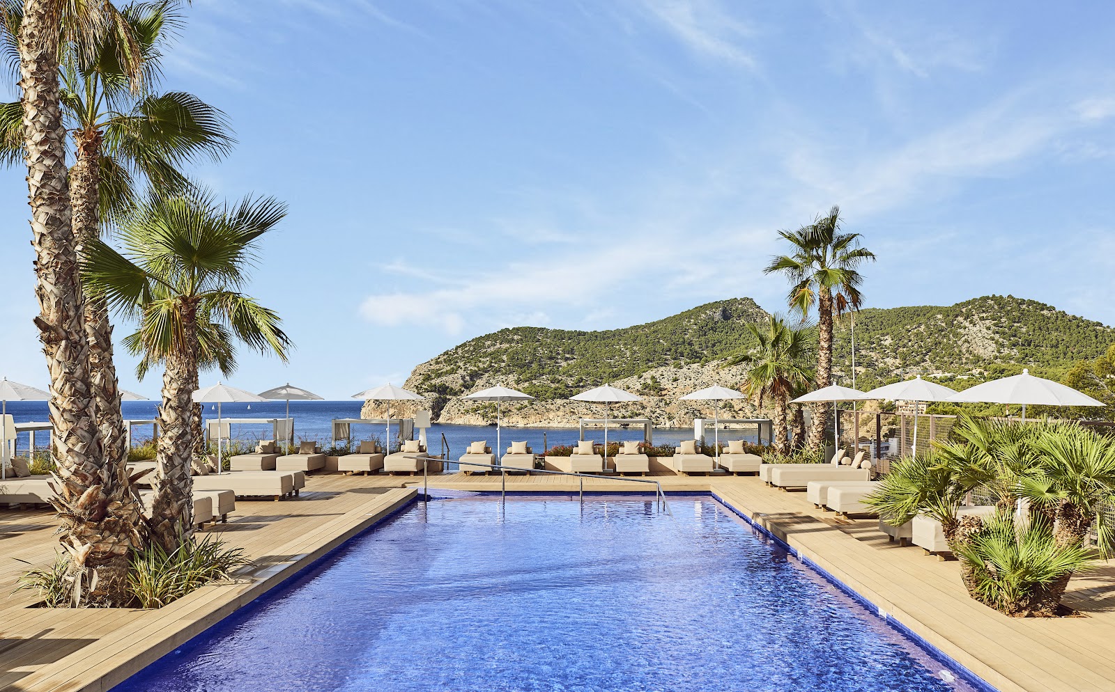 Resort pool with lounge chairs and white umbrellas overlooking a Mediterranean bay and forested hillside.