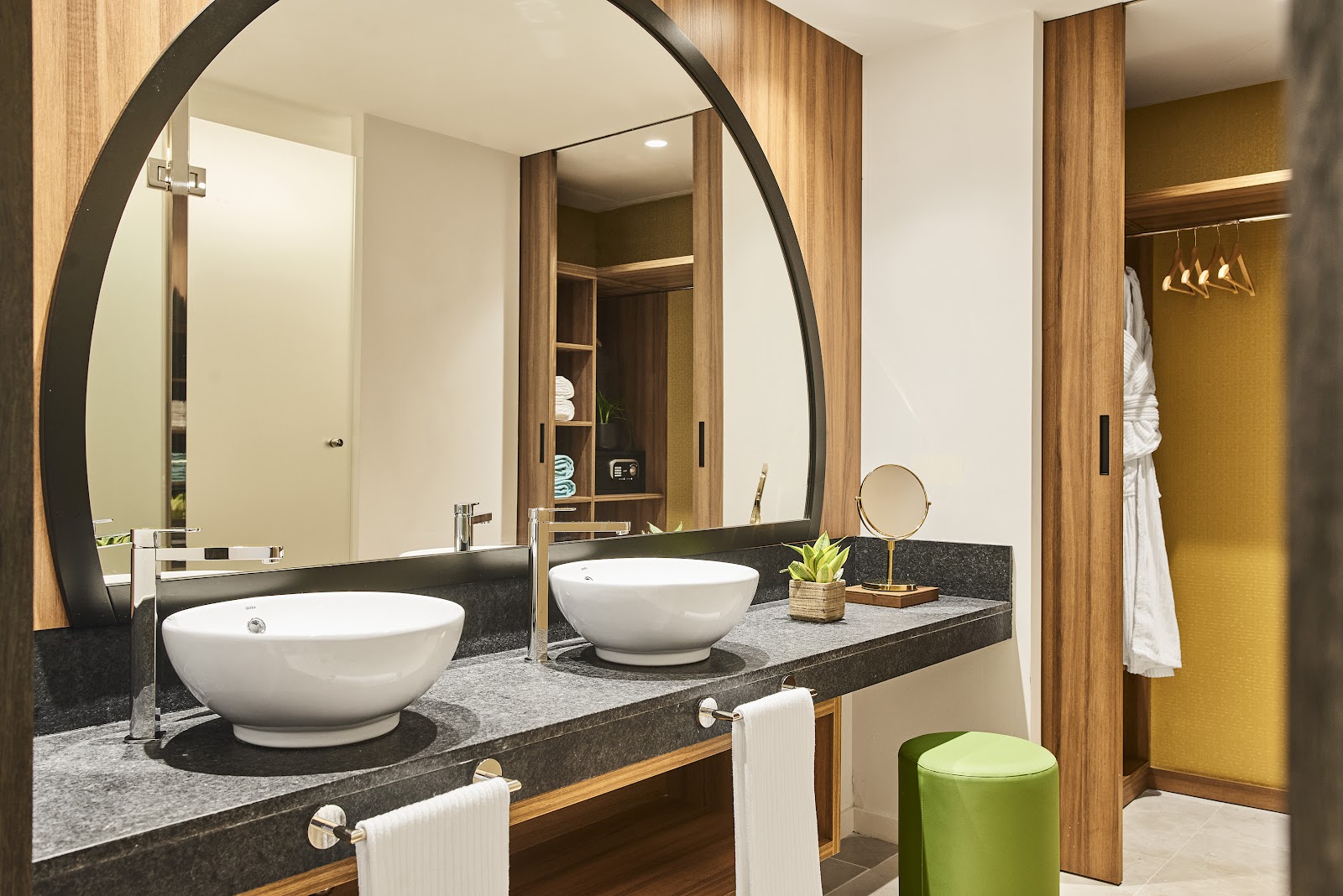 Bathroom with dual white vessel sinks on dark countertop, large curved mirror, wood paneling, and green accent stool at Zafiro Palace Andratx.