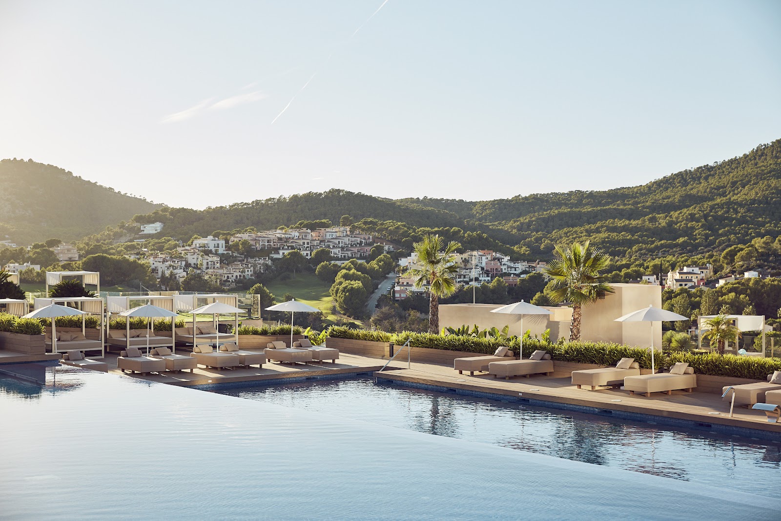 Resort pool with lounge chairs and white umbrellas overlooking forested hills and a coastal village in Mallorca.