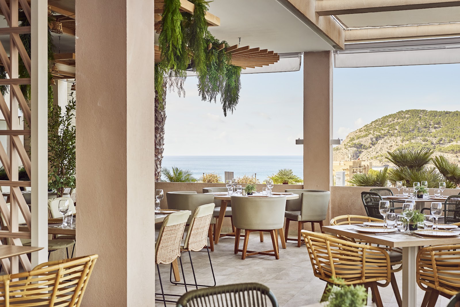 Open-air dining terrace at Zafiro Palace Andratx with round tables, woven chairs, and hanging greenery overlooking the Mediterranean Sea and forested coastline.