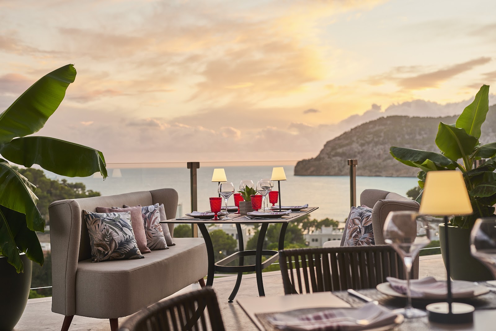 Outdoor dining terrace at Zafiro Palace Andratx with upholstered seating, set table, tropical plants, and bay view at sunset.