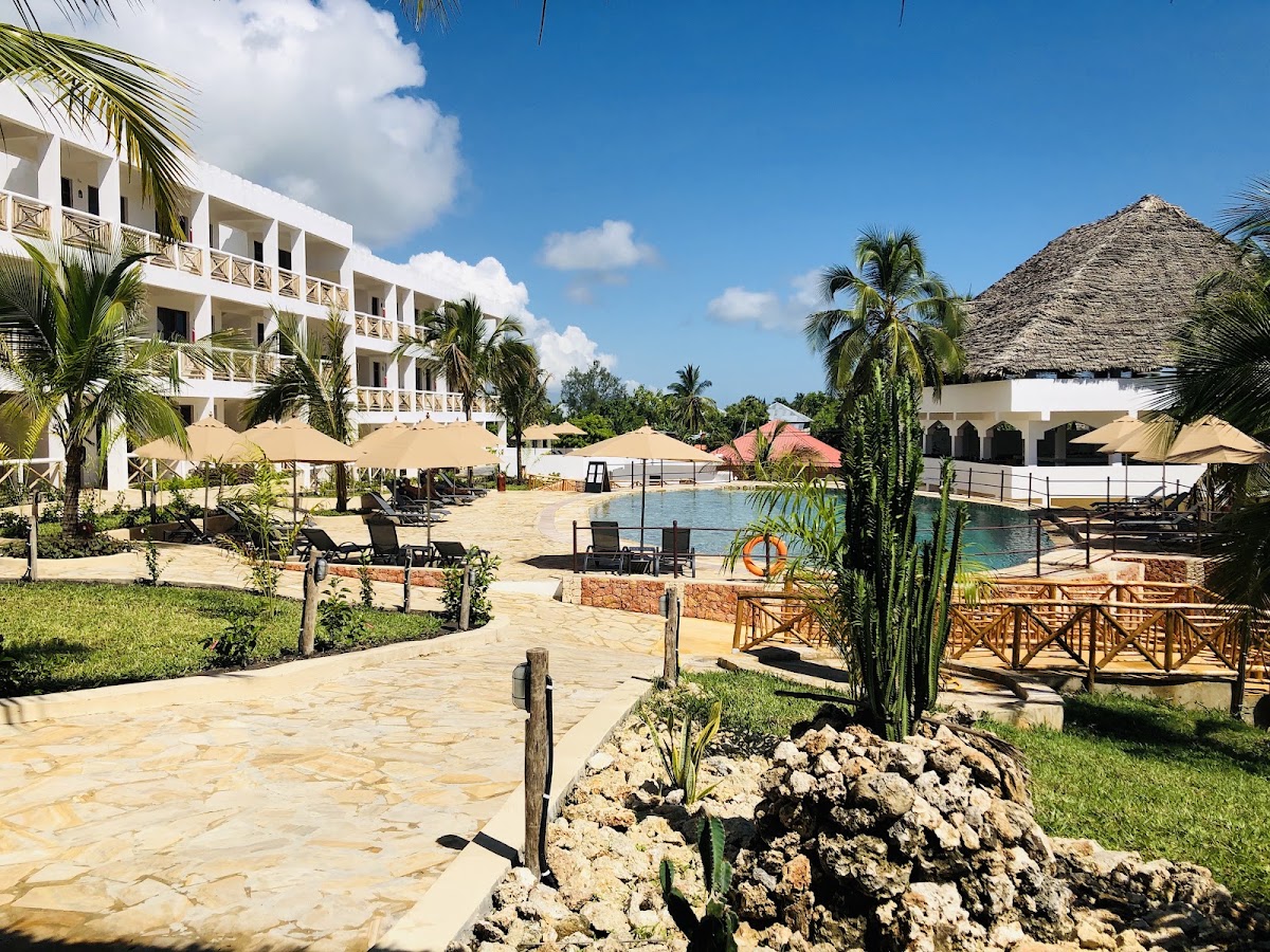 White multi-story resort building with palm trees, lounge chairs under tan umbrellas, and a lagoon pool with thatched-roof structures nearby under blue sky.