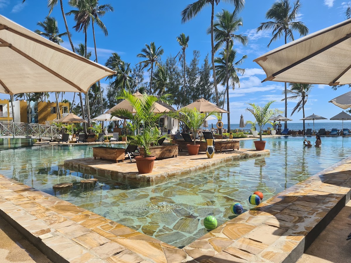 Zanzibar Bay Resort & Spa's lagoon pool with stone deck, palm trees, tan umbrellas, and ocean view under blue sky.