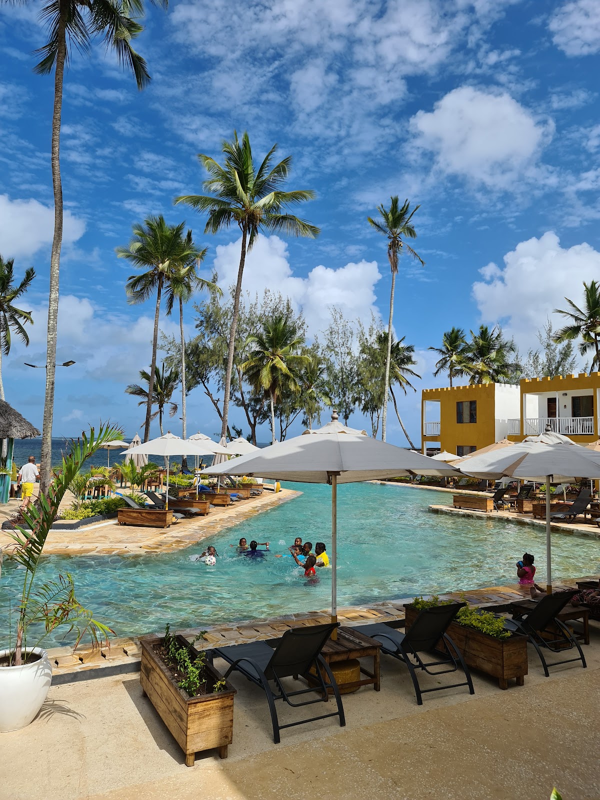 Guests swim in turquoise lagoon water at Zanzibar Bay Resort & Spa, with palm trees, beach loungers, umbrellas, and a yellow building visible.