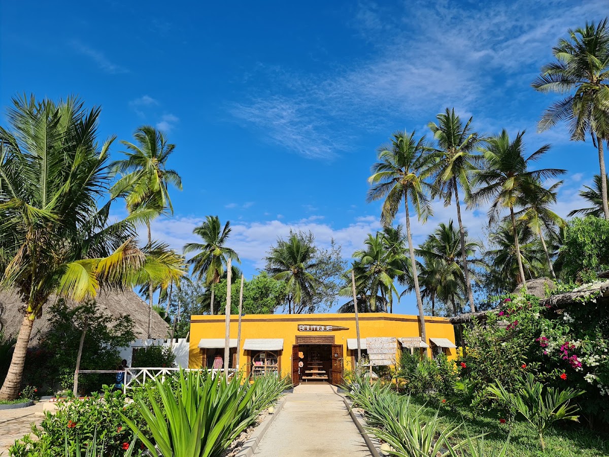 A bright yellow building with white awnings sits surrounded by tall coconut palms and tropical plants under a blue sky at Zanzibar Bay Resort & Spa.