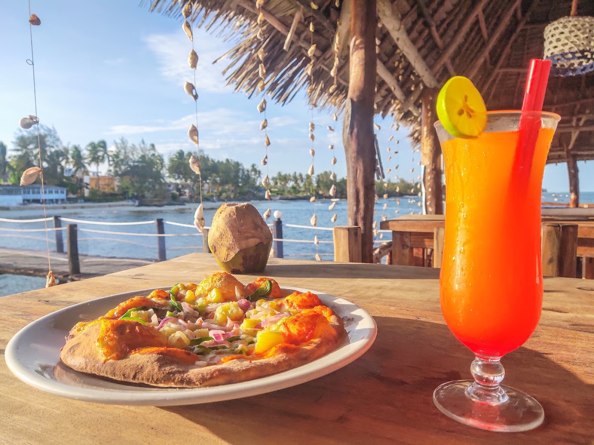 Pizza and tropical cocktail on wooden table under thatched roof with lagoon and palm trees visible behind.