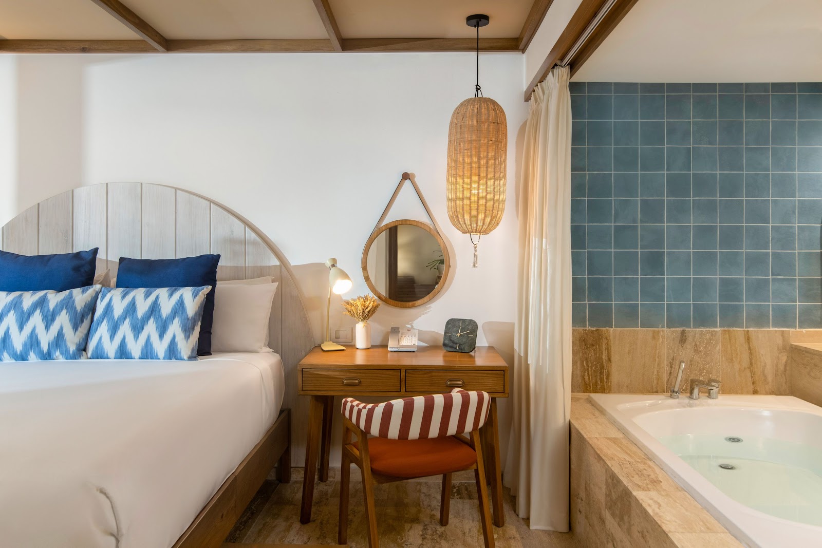 Resort guestroom with bed, wooden desk, woven pendant lamp, round mirror, and soaking tub with green tile wall at Zel Punta Cana.