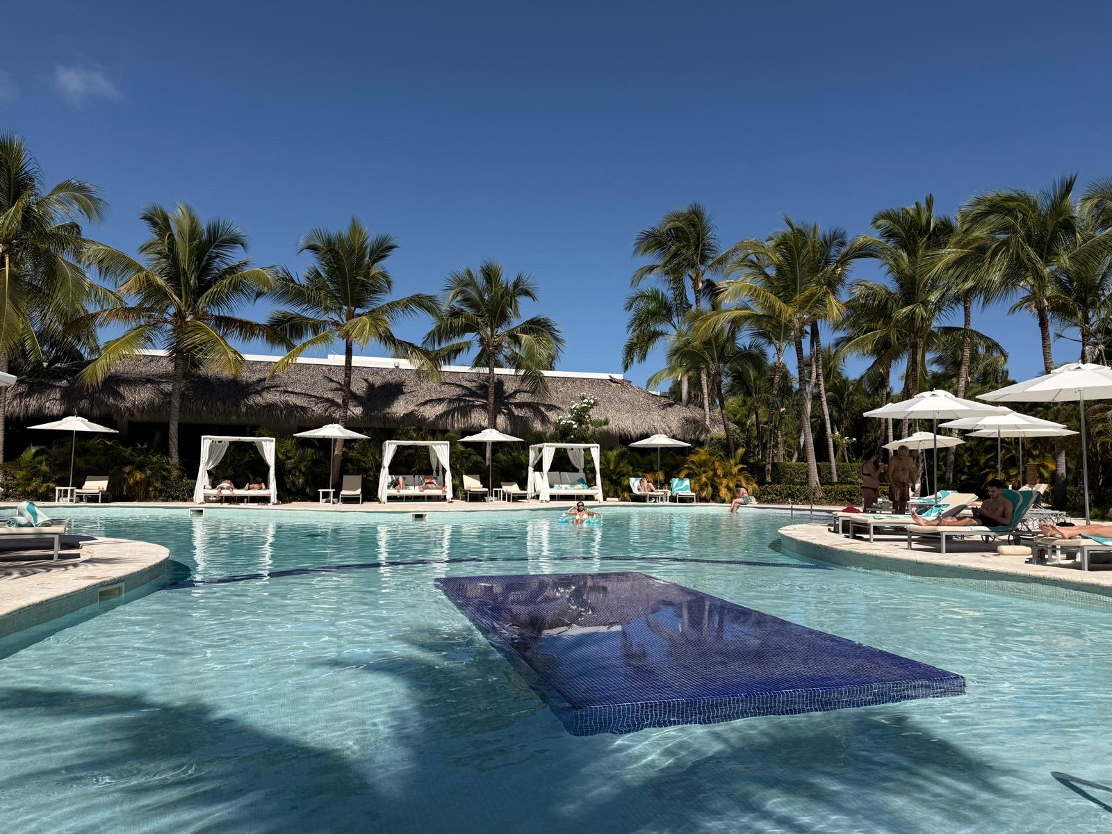 Resort pool with thatched-roof cabanas, white umbrellas, palm trees, and guests lounging under clear blue sky.