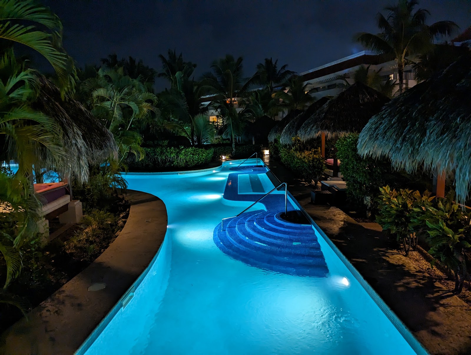 Glowing blue lazy river winds through palm trees and thatched palapas at night at Zel Punta Cana.