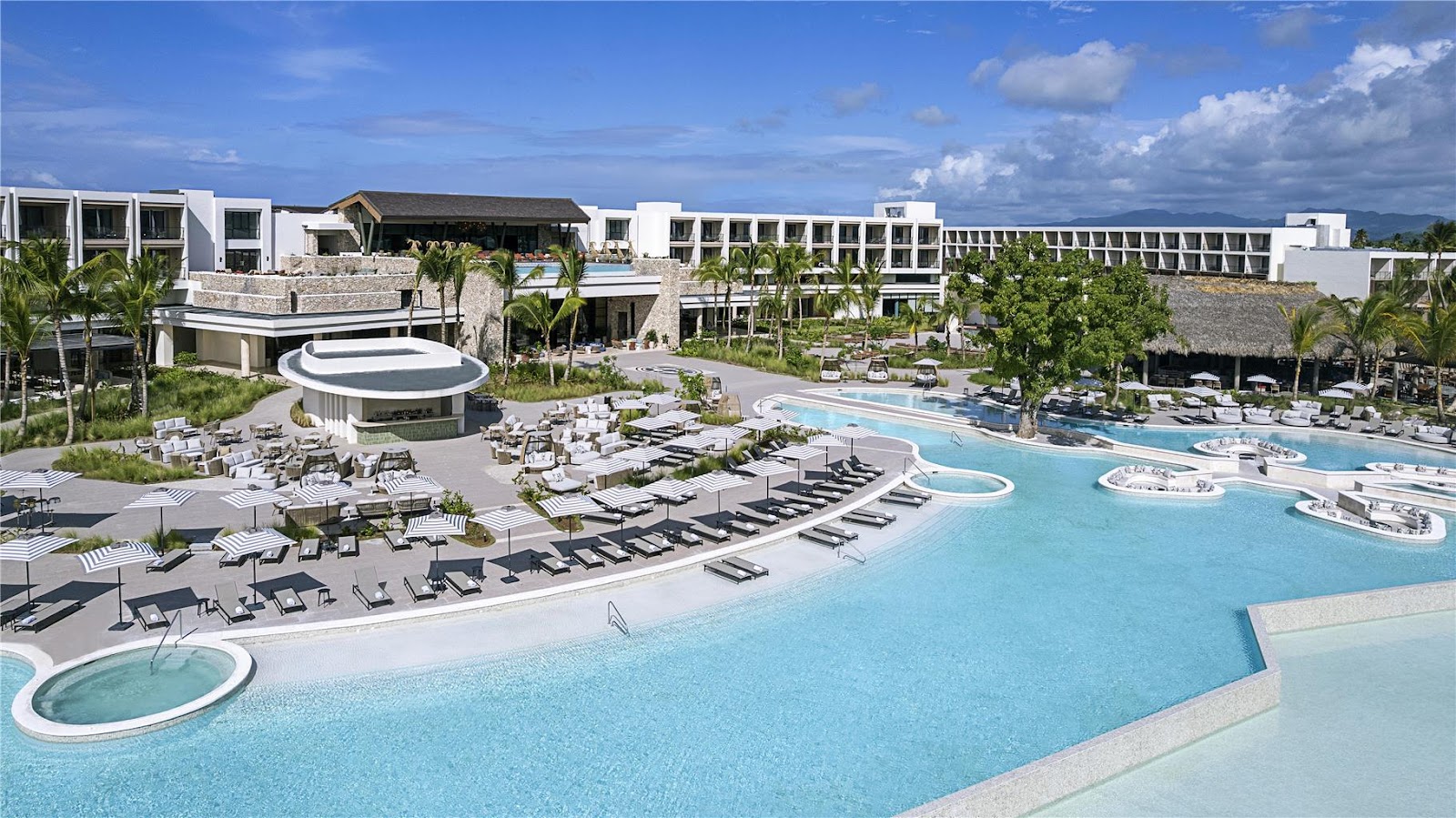 Expansive pool complex with turquoise waters, lounge chairs, and circular hot tubs surrounded by modern white resort buildings and palm trees under blue sky.