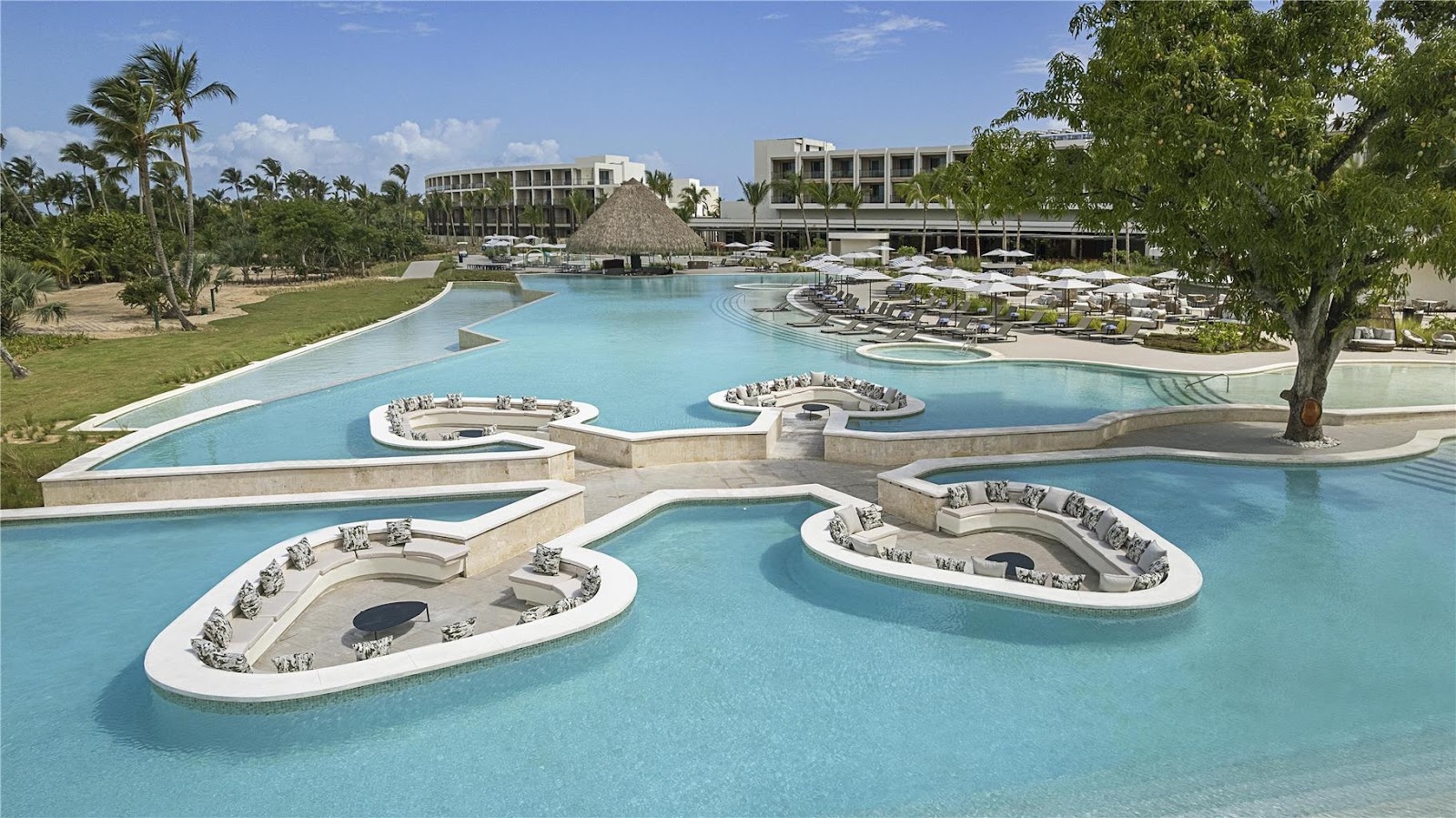 Resort pool complex with oval lounge islands, turquoise water, palms, and modern beige buildings under blue sky.