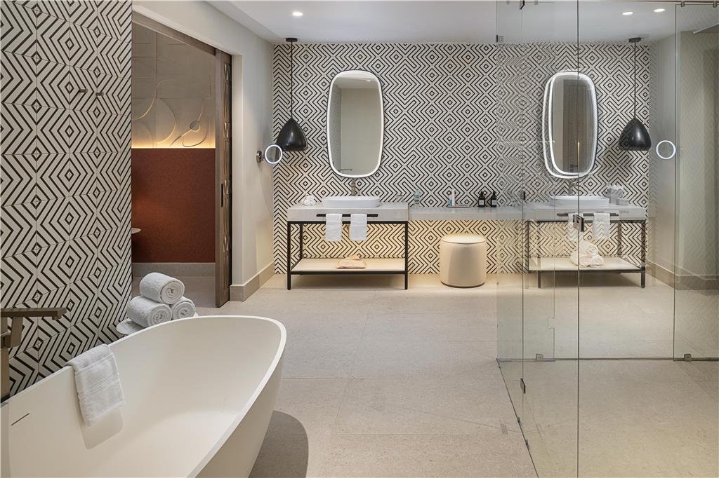 Modern spa bathroom with geometric tile walls, freestanding white tub, dual vanity with lit mirrors, and glass shower enclosure at Zemi Miches Punta Cana.