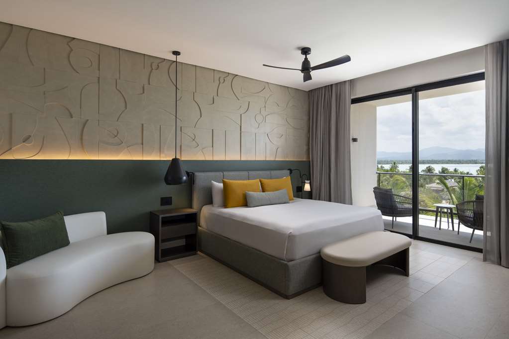 Modern resort bedroom with gray upholstered bed, curved white seating, and large balcony overlooking tropical landscape and water.