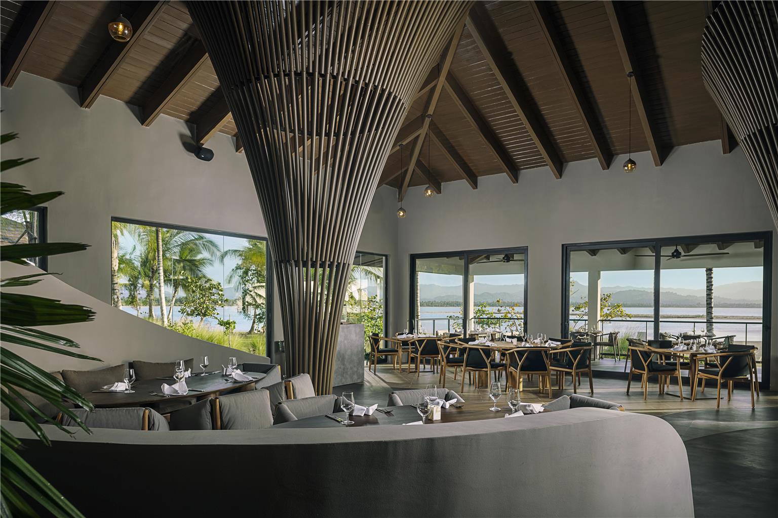 Modern dining hall at Zemi Miches Punta Cana with soaring wooden ceilings, gray upholstered seating, and floor-to-ceiling windows overlooking palm trees and the ocean.