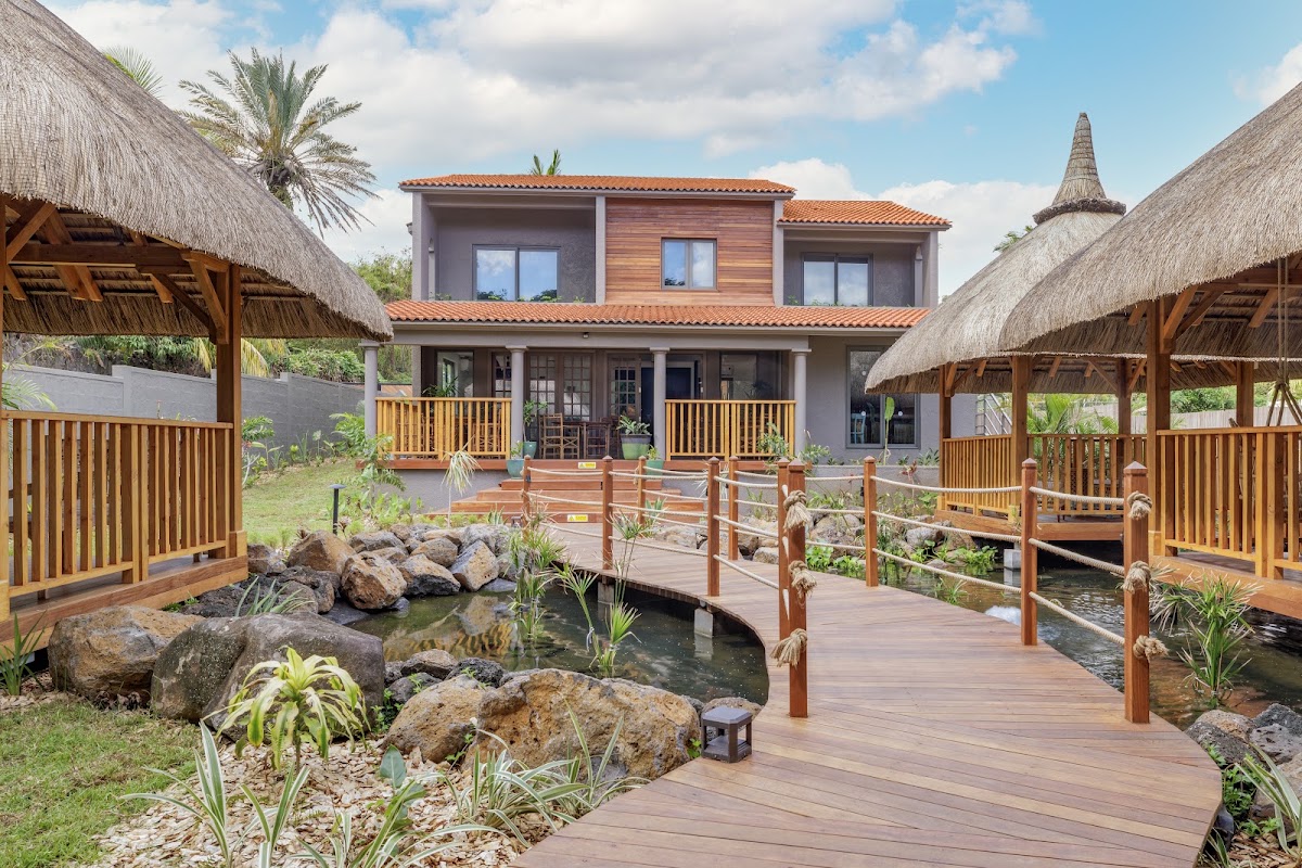 Wooden boardwalks cross shallow ponds with boulders and tropical plants leading to a two-story villa with thatched-roof bungalows on either side at Zilwa Attitude.