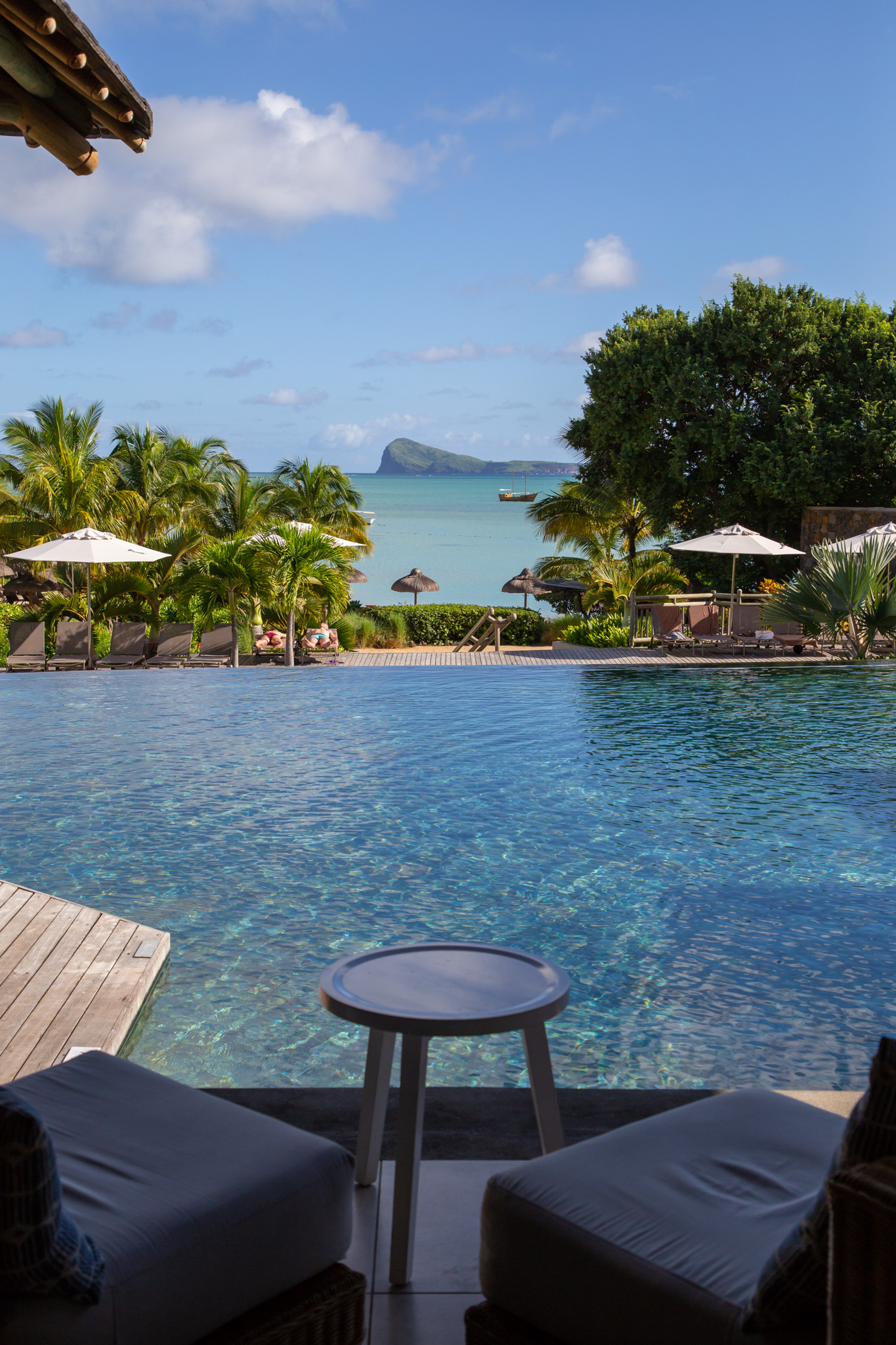 Lounge chairs and a small table overlook the lagoon pool at Zilwa Attitude, with palm trees, beach umbrellas, and a volcanic island visible across turquoise waters.