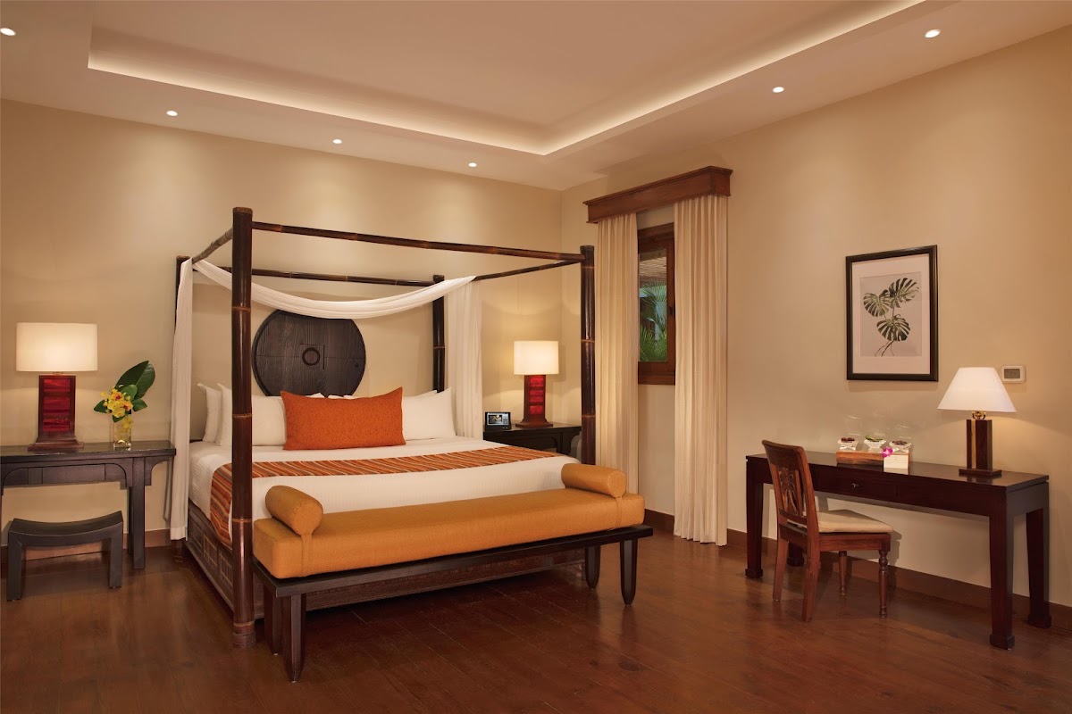 Spacious resort bedroom with four-poster canopy bed, orange accent pillow and bench, dark wood furniture, and warm lighting throughout.