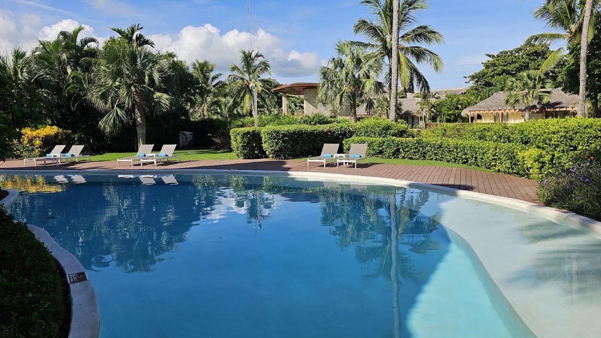 Large resort pool with blue water and wooden deck, surrounded by palm trees, lounge chairs, and thatched-roof buildings under partly cloudy sky.