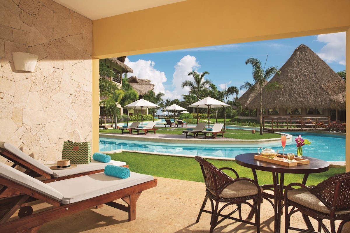 Open-air terrace with lounge chair and dining table overlooking curved pool, lawn, palm trees, and thatched-roof structures at Zoëtry Agua Punta Cana.