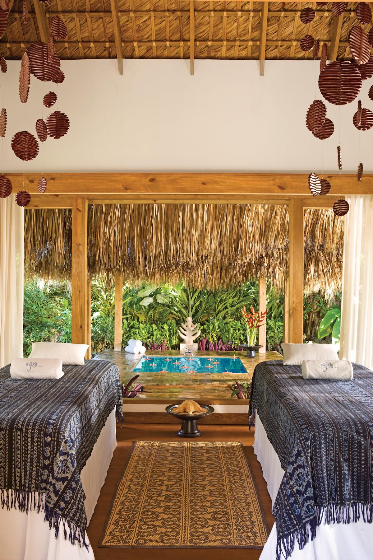 Spa treatment room with two massage beds facing large windows overlooking tropical gardens, thatched roof with hanging woven decorations.