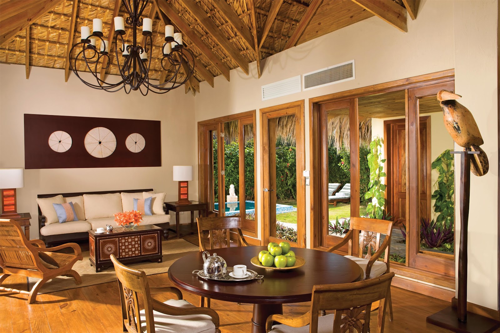 Beachfront dining pavilion with thatched roof, round tables dressed in white linens, and ocean view beyond palm trees and tiki structures.